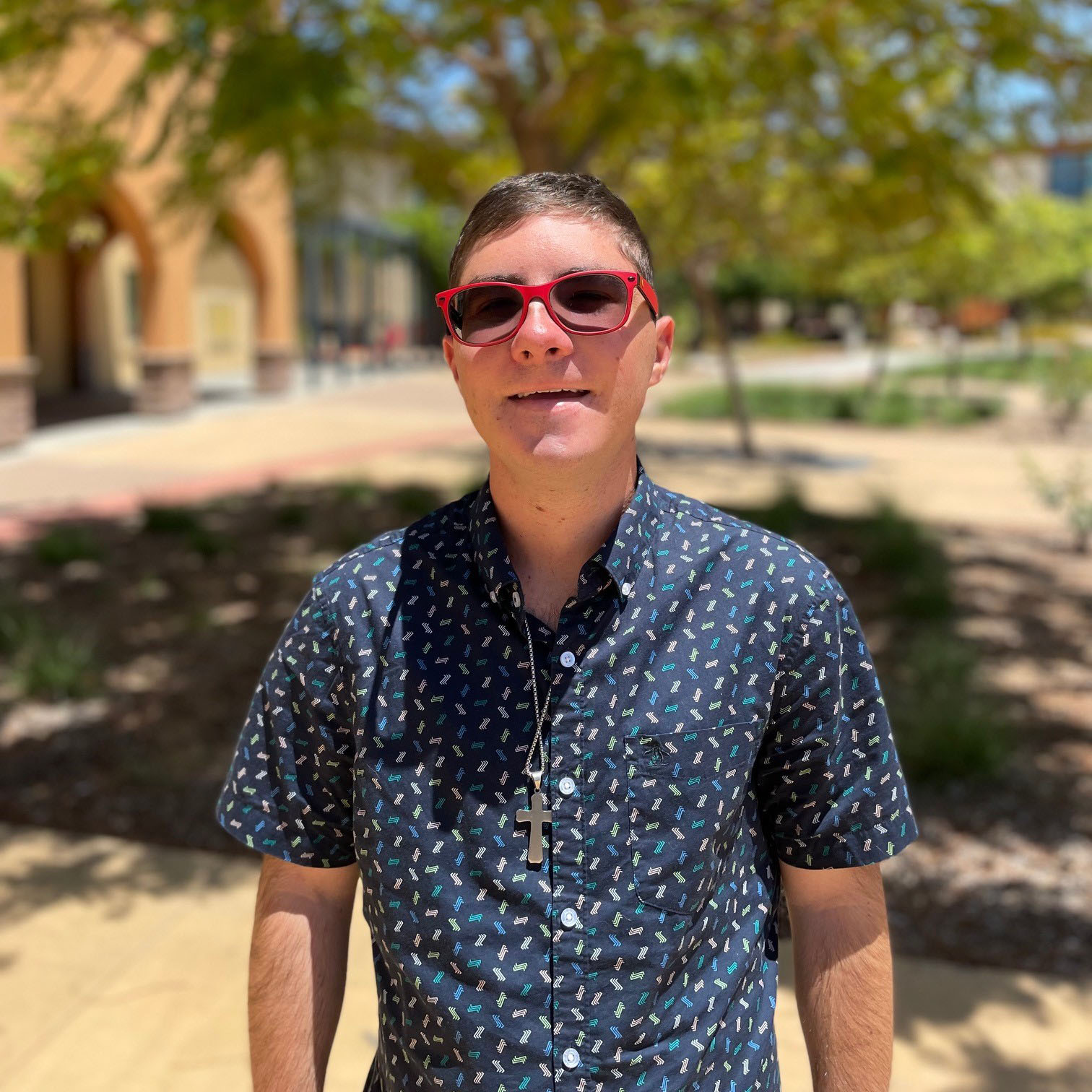 James Walker is standing outside on Miramar campus. He’s wearing a blue shirt and red glasses and smiling at the camera.