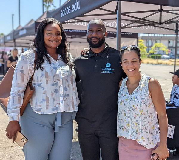 Three employees stand near an outdoor booth at a fair